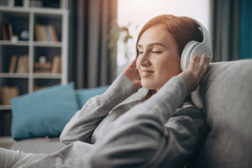 Relaxed young lady sitting on grey sofa with closed eyes and listening music in wireless headphones. Concept of enjoyment and leisure time