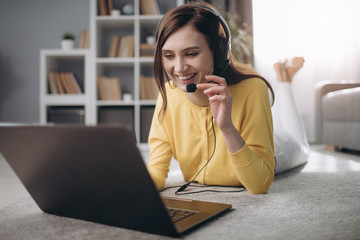 Smiling young woman in headset having video chat on laptop while lying on cozy carpet at home. Concept of freelance and online conversation