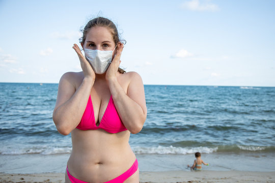 concept of fear of disease while traveling. young caucasian girl in a medical mask and in a swimsuit fearfully looks at the camera. In the background, on the beach by the sea, a child plays in blur.