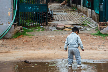 Indian lonely toddler boy walks into a puddle filled with rain water during monsoon season. Poor child on dirty streets walking alone in rainy weather in a poverty stricken slum area in Delhi, India.