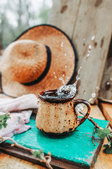 A splash of water in a brown ceramic mug on a green wood, with a cotton country towel, a currant leaf against and straw hat on background, a splash in the mug. tea party in nature