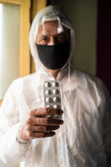 Senior man with corona preventive mask and PPE suit showing medicine strip in front of a window. Covid, Lock down and Home isolation