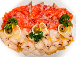 Sliced red salmon and white oily fish decorated with lemon wedges, dill sprigs and green olives on a white ceramic plate close-up