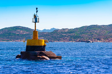 A view of a yellow and black nautical signaling buoy at sea in the blue sea on a sunny day with the coast of the island in the background, in Sardinia Italy