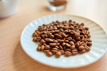 A white round plate with a handful of coffee beans is standing on the table. Selective focus