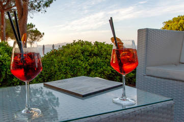A view of two fresh cocktails positioned spaced on a table of an empty outdoor bar, in the background a park on the sea and with the warm light of the evening sun, in Sardinia Italy