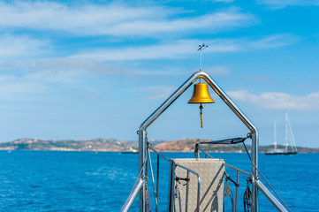 A view of the bell on the stern of the ship in the background blurred the blue sea the coast and some boats that leave the island towards the open sea on a sunny summer day, in Sardinia Italy
