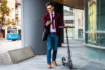 Smiling businessman with electric scooter standing in front of modern business building looking at phone. © Zoran Zeremski