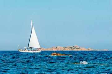 A view of a lonely white sailboat while sailing in the Mediterranean sea with a bird watching it and a small island in the background on a sunny day with blue sky on summer, in Sardinia Italy
