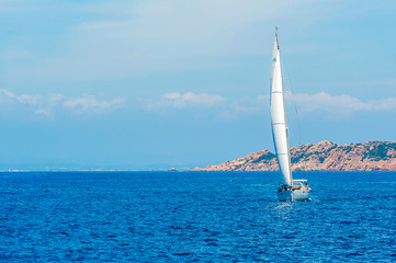 A view of a sailboat while sailing pushed by wind in the Mediterranean sea with the coast in the background on a day with the sun on summer, in Sardinia Italy
