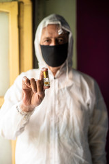 Senior man with corona virus preventive mask and PPE suit showing medicine ampul or vaccine in front of a window. Covid, Lock down and Home isolation