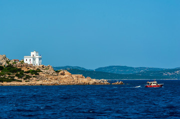 A view of a tourist boat with in the background a lighthouse on the rocks that is alluring from the coast to visit the islands of the archipelago on a sunny day in summer, in Sardinia Italy
