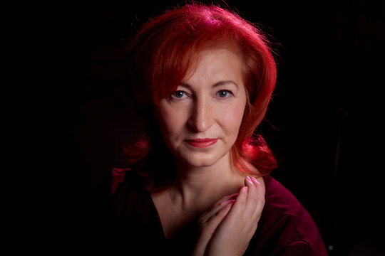 Stylish elderly woman with red hair in vinous dress posing in studio with dark background and red flash and light
