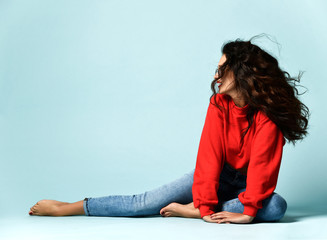 Curly brunette woman in smoked blue glasses, red sweater and blue jeans sits posing on the floor...