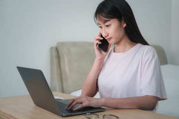 Asian women working at home are using laptop and mobile phone to serious work in the bedroom on the early morning.