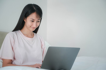 Happy casual beautiful asian woman working on a laptop sitting on the bed in the house.