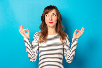 Portrait of young friendly woman with a smile in a casual t-shirt making a zen gesture and looking at the stern on isolated blue background. Emotional face. Concept of relaxation, meditation, stress