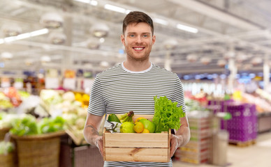 Fototapeta premium sustainability, food shopping and eco friendly concept - happy smiling man with fruits and vegetables in wooden box over eco market or grocery store on background