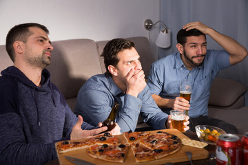 Three anxious men watching tv, drinking beer at home