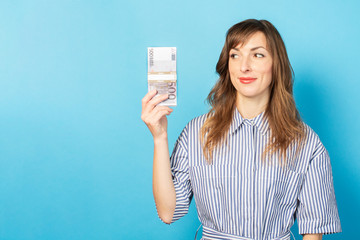 Portrait of a young friendly woman with a smile in a dress holding a bundle of money in her hands and looking at her on an isolated blue background. Emotional face. Concept of wealth, win, credit