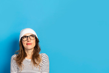 Portrait of a Cute Young woman with glasses, a hat and a jacket looks up with a happy face on a blue background. Emotional face. Gesture to think, dream, plan
