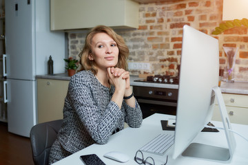 A woman smiles and works remotely on a desktop computer in her studio. A lady sits arms crossed during a video conference at home. A female professor listening to student's answers on an online lesson