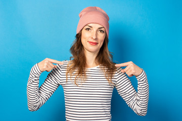 Cute young woman in a hat and striped sweater points her fingers at herself on a blue background....