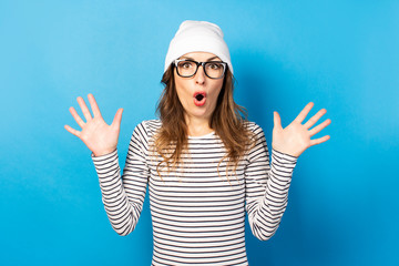 Portrait Cute young woman in glasses, hat and jacket spreads her arms with a surprised face on a blue background. Emotional face. Gesture shock, surprise