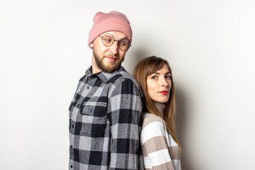 Young man with a beard in a hat and a plaid shirt and a girl in a sweater are standing back to back on an isolated light background. Man looks at a woman. Emotional face