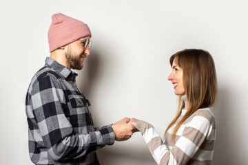 Young man with a beard in a hat and a plaid shirt and a girl in a sweater hold hands on an isolated light background. Emotional face. Happy couple dating