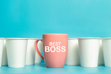 Pink cup surrounded by white paper cups on a blue background. Concept boss, unique, friendly team, feminism. Copy space. Added text Best Boss
