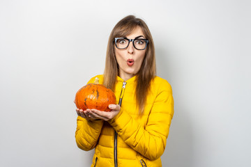 young girl in a yellow jacket holds a pumpkin with a frightened face on a light background. Halloween concept, autumn, celebration