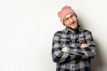 Portrait of a young man with a beard in a hat, plaid shirt and glasses with arms crossed on his chest smiling on an isolated light background. Emotional face