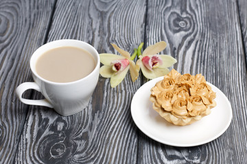 Cupcake and a cup of coffee with milk. Nearby are orchid flowers. Cake decorated with flowers made of butter cream. The cream has a caramel color. On brushed pine boards painted black.