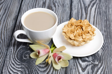 Cupcake and a cup of coffee with milk. Nearby are orchid flowers. Cake decorated with flowers made of butter cream. The cream has a caramel color. On brushed pine boards painted black.