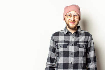 Naklejka premium Portrait of a young man with a beard in a hat, plaid shirt and glasses smiling on an isolated light background. Emotional face