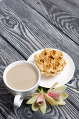 Cupcake and a cup of coffee with milk. Nearby are orchid flowers. Cake decorated with flowers made of butter cream. The cream has a caramel color. On brushed pine boards painted black.