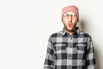 Portrait of a young man with a beard in a hat, plaid shirt and glasses looks to the side with a surprised face on an isolated light background. Emotional face