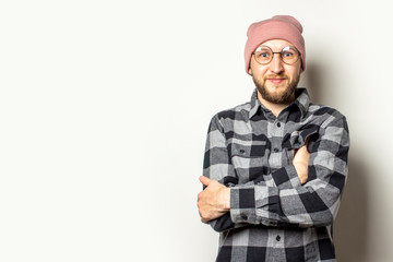 Portrait of a young man with a beard in a hat, a plaid shirt and glasses with arms crossed on his chest on an isolated light background. Emotional face