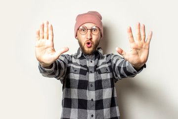 Naklejka premium Portrait of a young man with a beard in a hat, a plaid shirt stretched out his hands in front of him with his palms to the viewer on an isolated light background. Emotional face. Stop gesture, enough