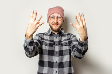 Naklejka premium Portrait of a young man with a beard in a hat, a checkered shirt extended his arms in front of him with the back of his hand on an isolated light background. Emotional face. Stop gesture, enough