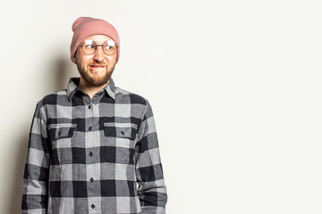 Portrait of a young man with a beard in a hat, a plaid shirt and glasses looks away with an embarrassed face on an isolated light background. Emotional face