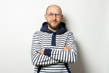 Portrait of a Bald Young man with a beard in glasses of a sweater with a hood crossed his arms over his chest on an isolated light background. Emotional face. Concentrated gesture