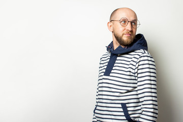 Portrait of a Bald Young man with a beard in glasses, a sweater with a hood looks back on an isolated light background. Emotional face. Gesture looking back