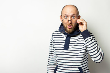 Portrait of a Bald Young man with a beard, a sweater with a hood lowered his glasses on his nose with a surprised face on an isolated light background. Emotional face. Copy space