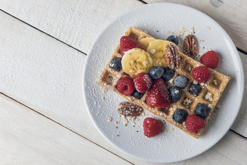 Waffle with banana slices, oat, pecans and berries served on a white plate over a white rustic wooden table. From above with copy space.