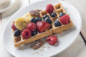 Homemade waffle topped with banana slices, oat, pecans and berries served on a white plate over a white rustic wooden table. Closeup.