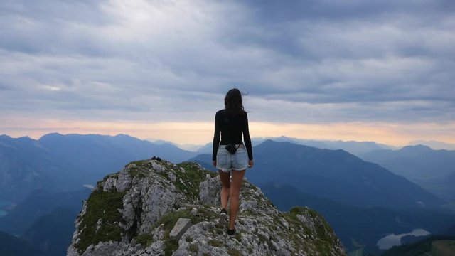 Young pretty hiker girl walking at mountain peak Schafberg Austria. 60fps slowmo footage. Sexy woman with camera climbing in alpine scenery. Sunrise at summit and clouds dramatic epic mood