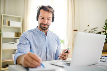 Mature businessman with headphones and laptop indoors in home office, working.