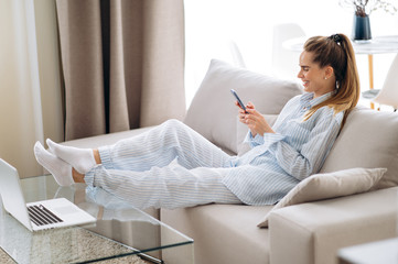 Chatting online. Weekend mood. Young attractive girl in home clothes sits on her cozy sofa and...